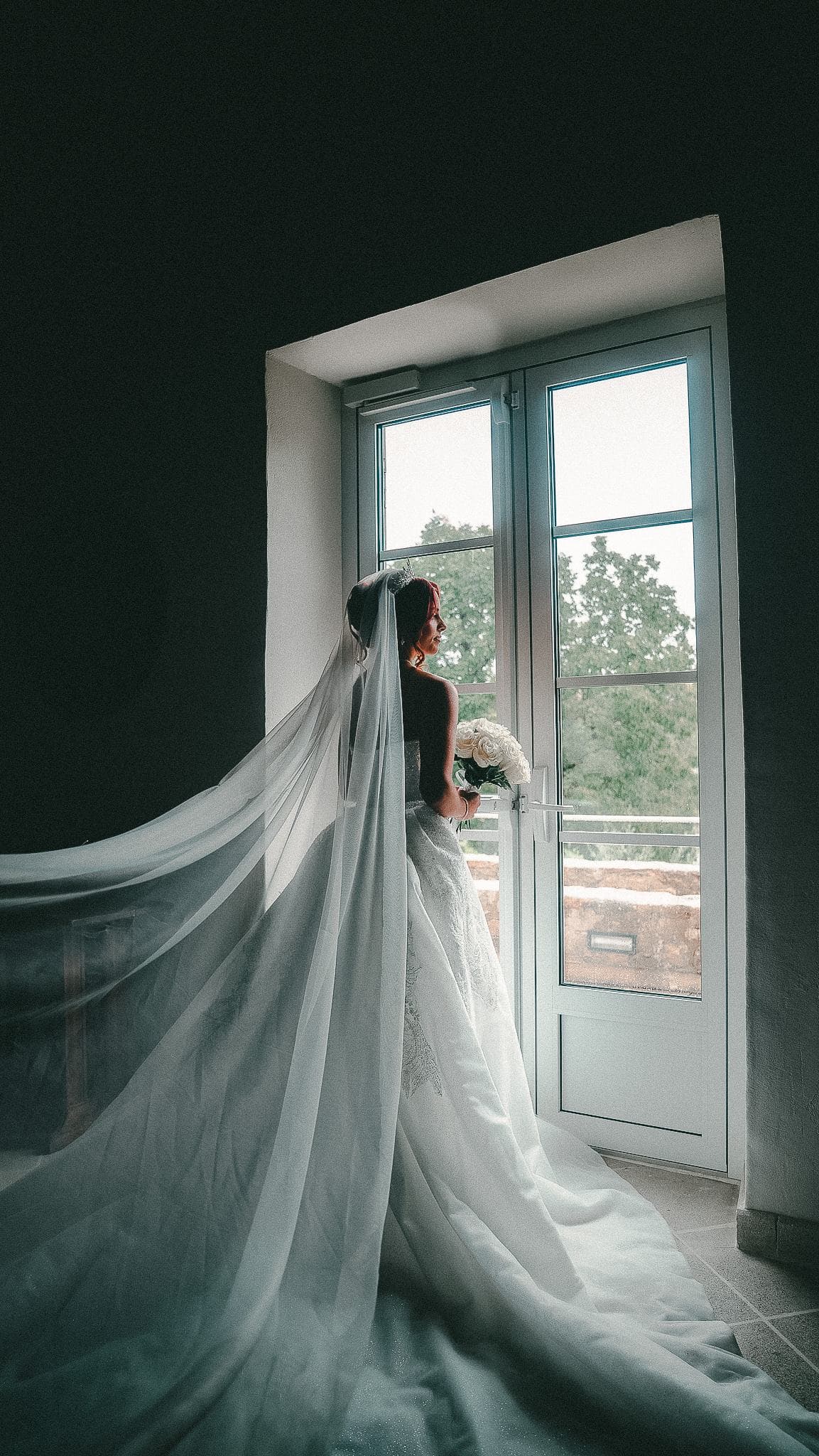 La mariée de dos devant une porte-fenêtre, son voile déployé dans la lumière