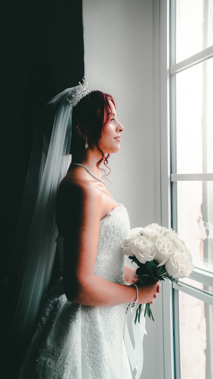 La mariée de profil devant une fenêtre, son bouquet de roses blanches à la main