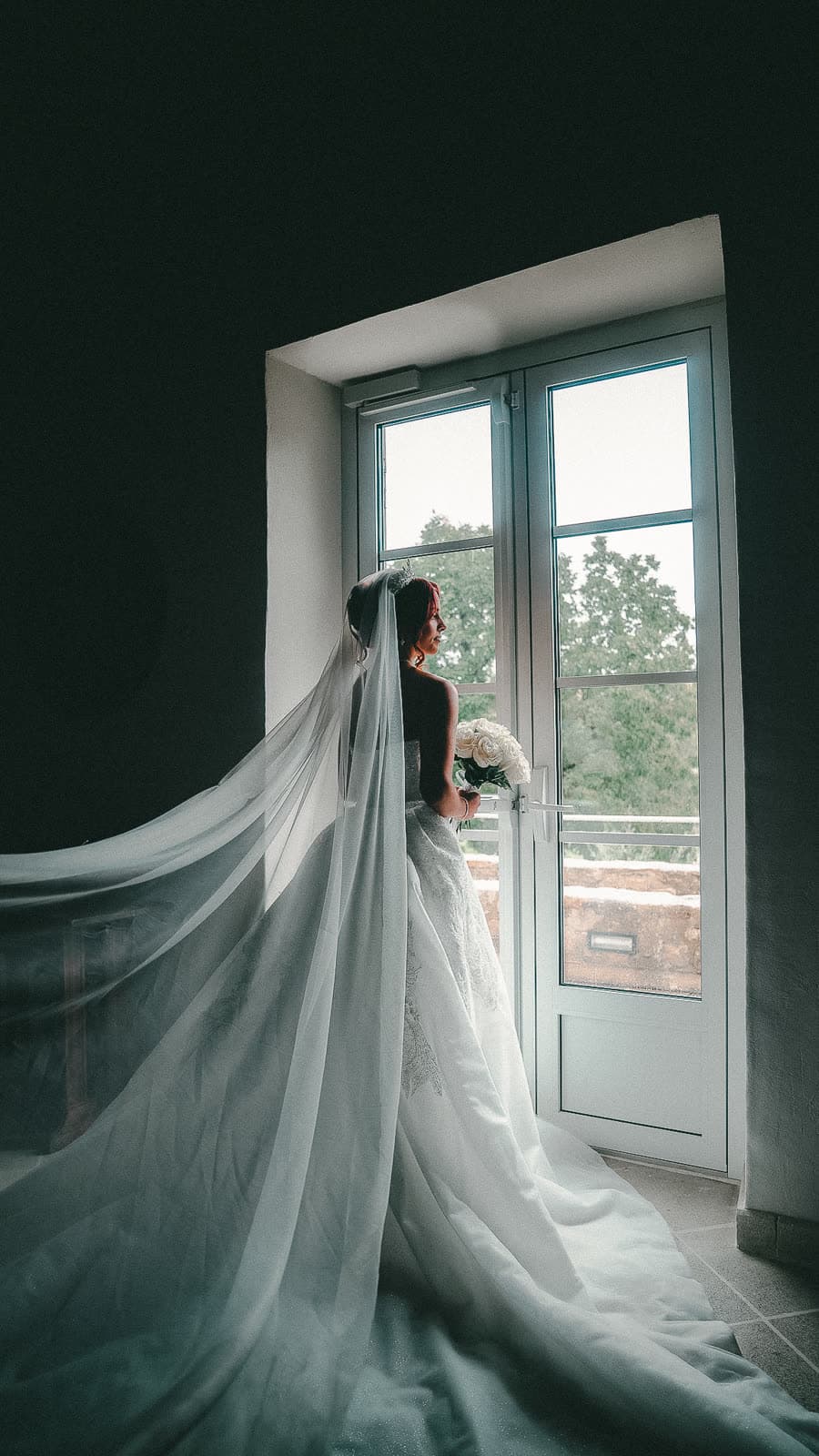 La mariée de dos devant une porte-fenêtre, sa traîne déployée